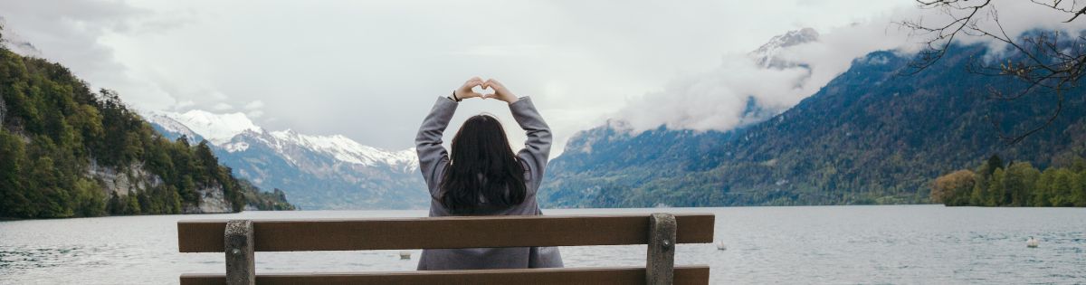 vrouw op parkbank met bergen die leren mediteren met liefde terwijl ze haar handen in een hartvorm boven haar hoofd houdt